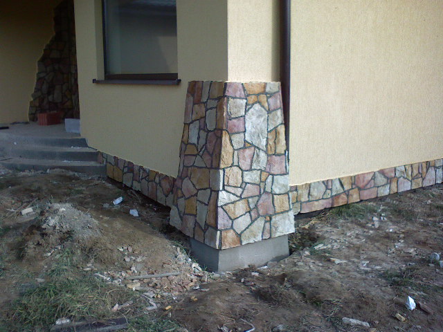 Wykończenie elewacji domu kamieniem dekoracyjnym, widoczny fragment ściany z oknem i narożny filar obłożony kamieniem w odcieniach brązu, beżu i szarości, w tle fragment schodów wejściowych.